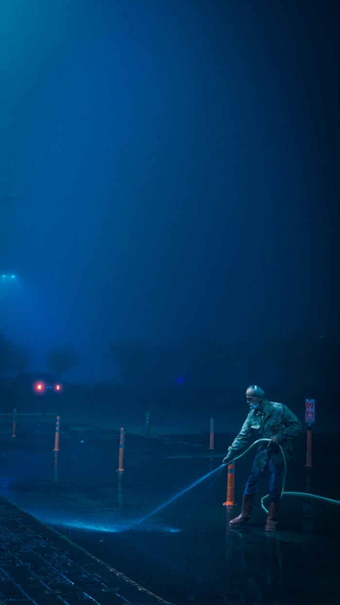 About A man using a water hose to clean a wet street at night under streetlights.